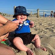 Dakeem is registered to the contest to win money with this photo: baby, beach, sand, bucket_hat, blue_shirt, nike_air, smiling, happy, barefoot, adult_hands, sunlight, outdoor, summer, fence, sky, person, child, summer_clothing, playful, vacation