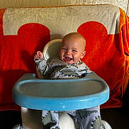 Colt is registered to the contest to win money with this photo: baby, child, infant, smiling, happy, dinosaur_pajamas, baby_seat, high_chair, blue_tray, orange_blanket, beige_blanket, indoor, cute, sitting, joyful, face, person, footwear, pajamas, young_child