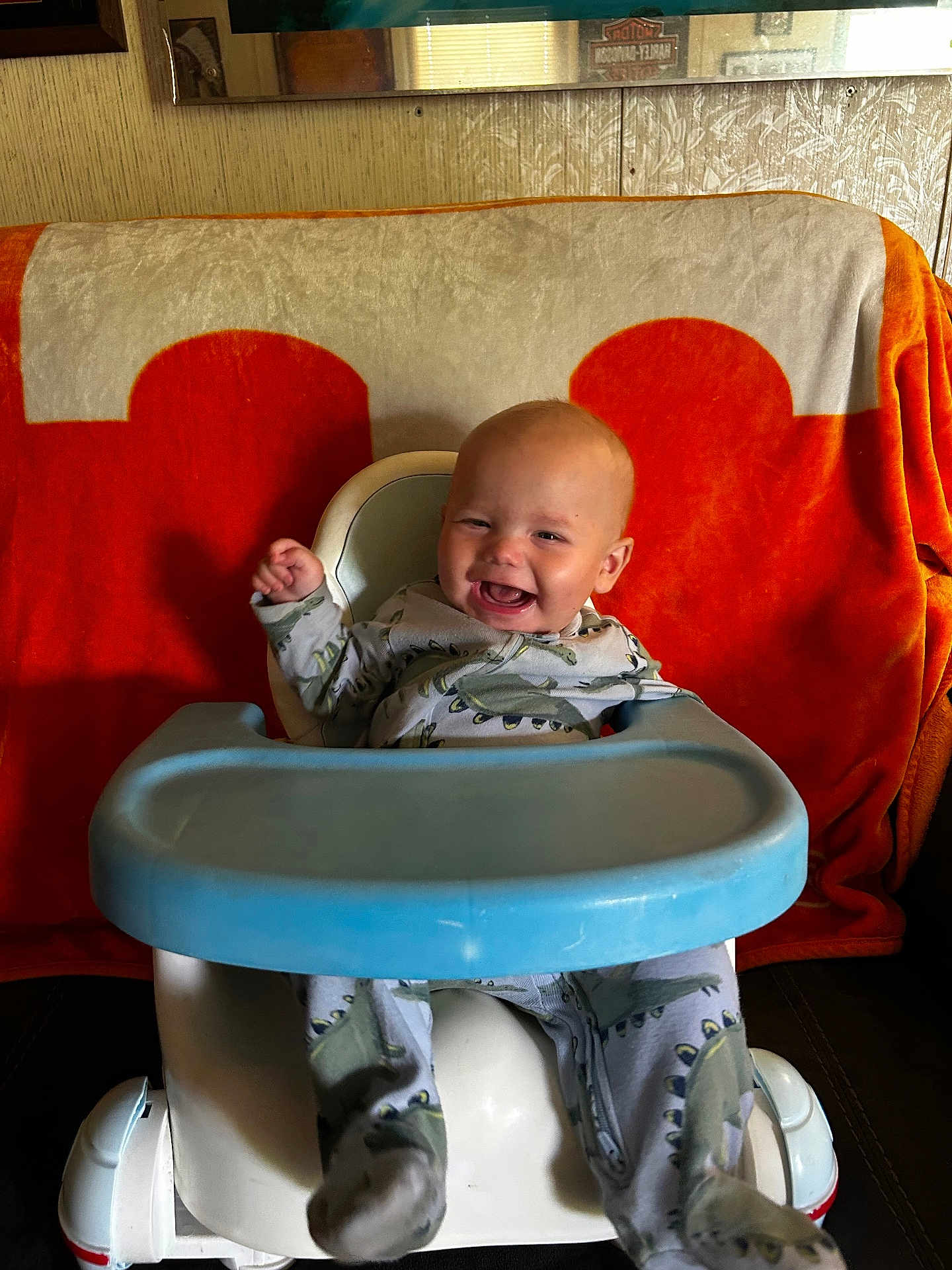 Colt is registered to the contest to win money with this photo: baby, child, infant, smiling, happy, dinosaur_pajamas, baby_seat, high_chair, blue_tray, orange_blanket, beige_blanket, indoor, cute, sitting, joyful, face, person, footwear, pajamas, young_child