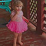 child, toddler, pink_tutu, barefoot, porch, wooden_floor, fence, outdoor, curly_hair, thoughtful, colorful_chair, green_chair, rustic, summer, daylight, person, young_child, standing, casual, cute