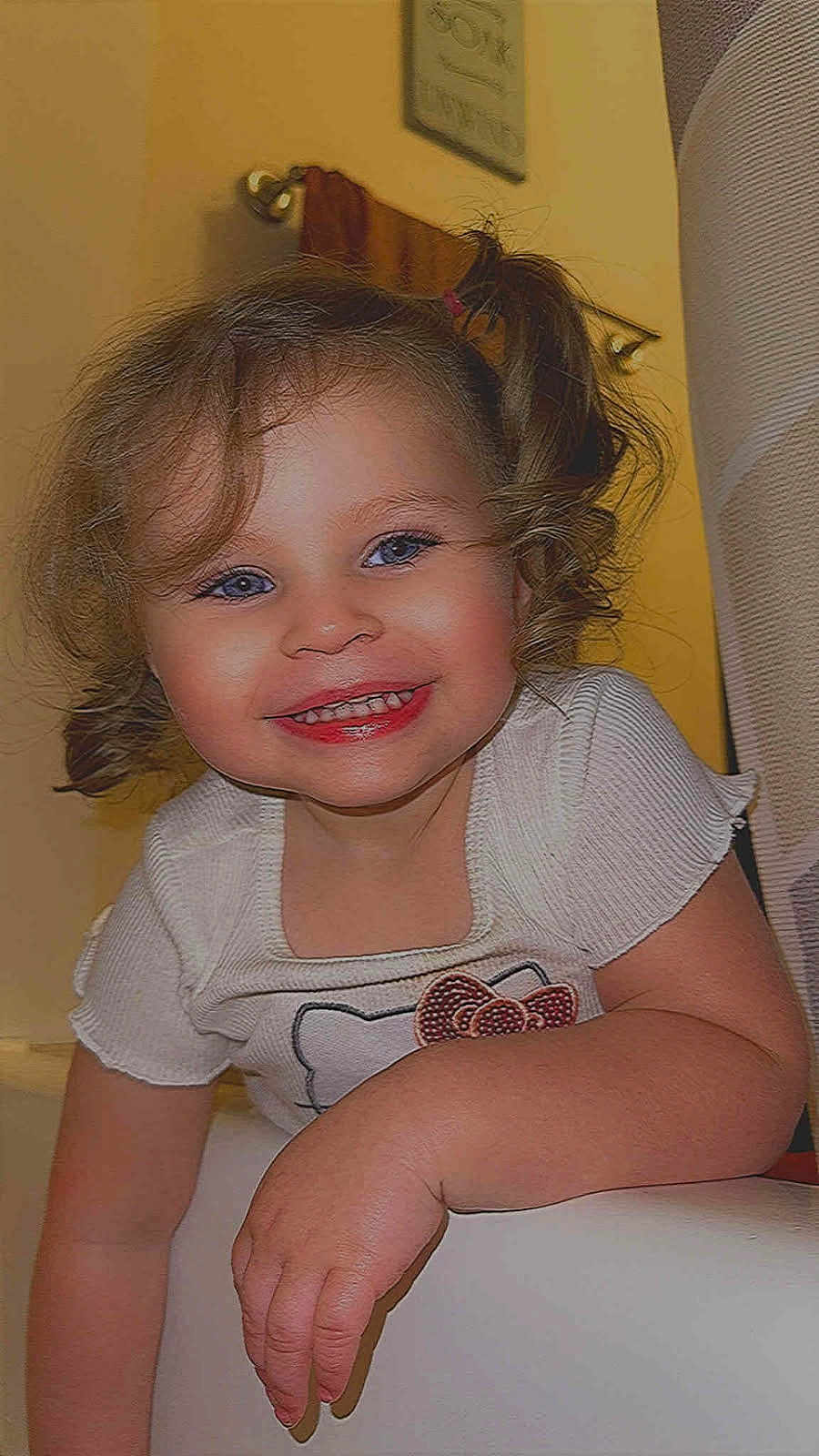 Violet is registered to the contest to win money with this photo: toddler, child, smile, curly_hair, pony_tail, white_shirt, indoor, yellow_wall, towel, framed_picture, hand, face, happy, cute, portrait, person, young_child, skin, baby, cheerful