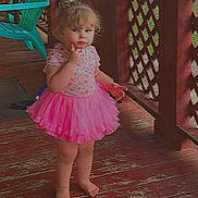 Violet is registered to the contest to win money with this photo: barefoot, casual_clothing, child, curly_hair, daylight, expression, fence, green_chair, lattice, outdoor, person, pink_tutu, playful, porch, standing, summer, thinking, toddler, wooden_floor, young_child