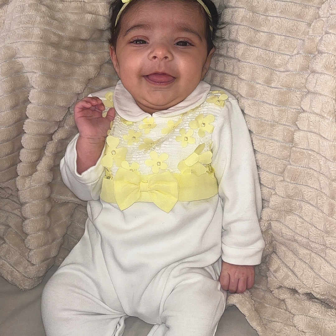 Le Ara is registered to the contest to win money with this photo: baby, blanket, child, cozy, cute, face, footed_pajamas, happy, headband, indoors, infant, lying_down, portrait, small_hand, smiling, soft_texture, toddler, white_outfit, yellow_bow, yellow_flowers