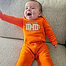 Kaden joined the competition — help win amazing prizes! baby, child, couch, cute, fabric, face, furniture, happy, head, indoor, infant, m_and_m, onesie, orange_clothing, person, playful, raising_hand, sitting, smiling, sofa