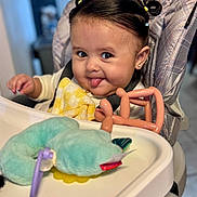 Kenzie is registered to the contest to win money with this photo: baby, high_chair, tongue_out, plush_toy, balloon_animal, yellow_blanket, hair_accessories, face, smile, child, indoor, cute, person, seat, clothing, earring, expression, toy, infant, happy
