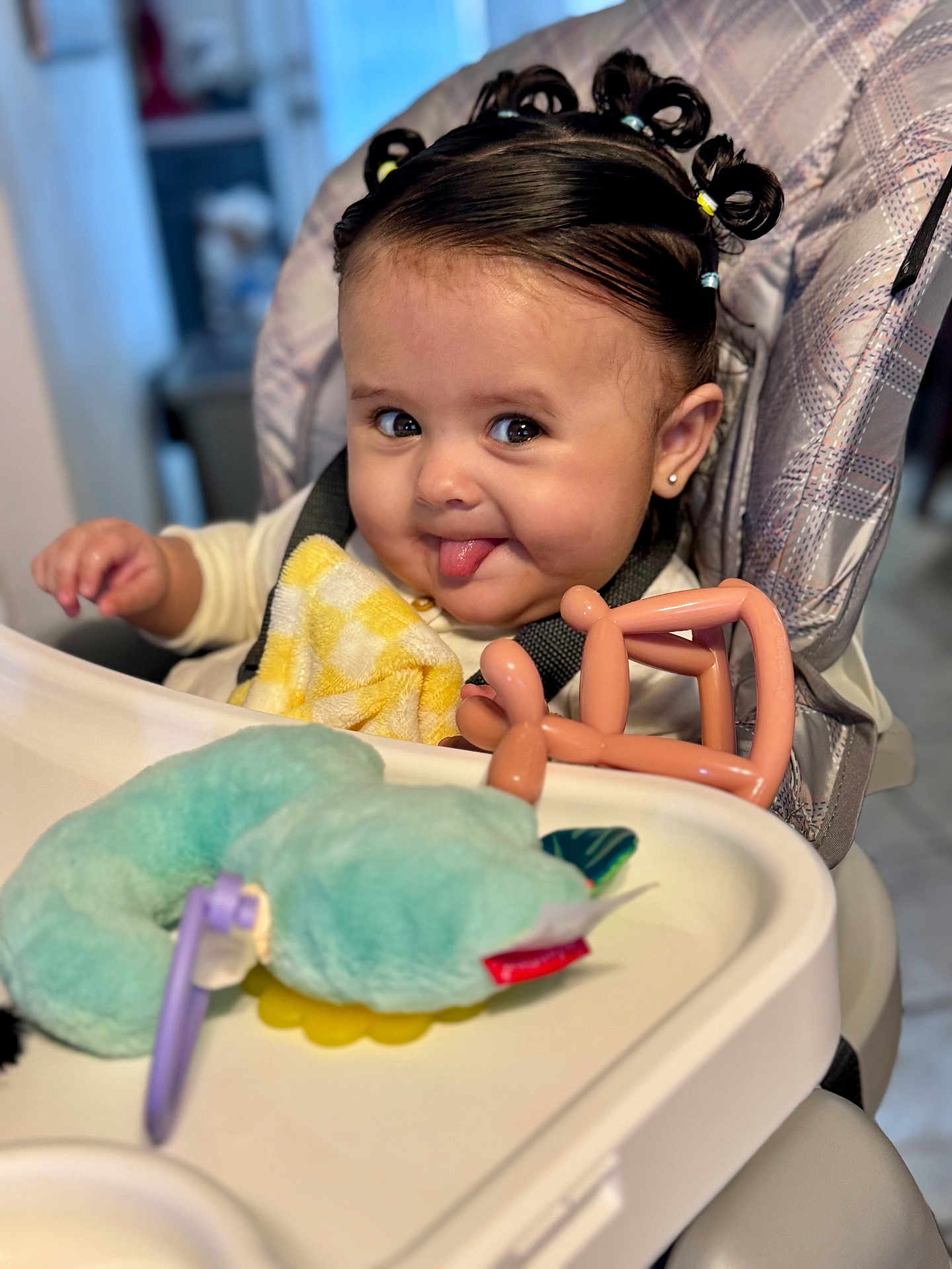 Kenzie is registered to the contest to win money with this photo: baby, high_chair, tongue_out, plush_toy, balloon_animal, yellow_blanket, hair_accessories, face, smile, child, indoor, cute, person, seat, clothing, earring, expression, toy, infant, happy