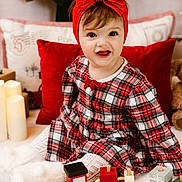 Mia is registered to the contest to win money with this photo: toddler, child, red_bow, plaid_dress, toy_train, candles, pillows, holiday, indoors, smiling, happy, cute, warm_lighting, cozy, white_boots, headband, playful, wooden_toy, festive, portrait