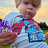 toddler, child, flower, purple_flower, spiderman, shirt, blonde_hair, blue_eyes, outdoor, sunset, green_field, nature, portrait, hand, grass, sky, casual_clothing, person, cute, young