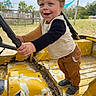 Nolan is registered to the contest to win money with this photo: toddler, child, boy, blue_eyes, steering_wheel, yellow_vehicle, outdoor, grass, trees, rustic, play, clothing, pants, sneakers, smile, happy, portrait, daylight, fence, nature