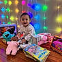 Maelys is registered to the contest to win money with this photo: baby, smiling, toys, plush_doll, book, wooden_floor, colorful_lights, fairy_lights, indoor, child, playtime, happy, pink, blue, packaging, music_player, floor, wall, person, cute