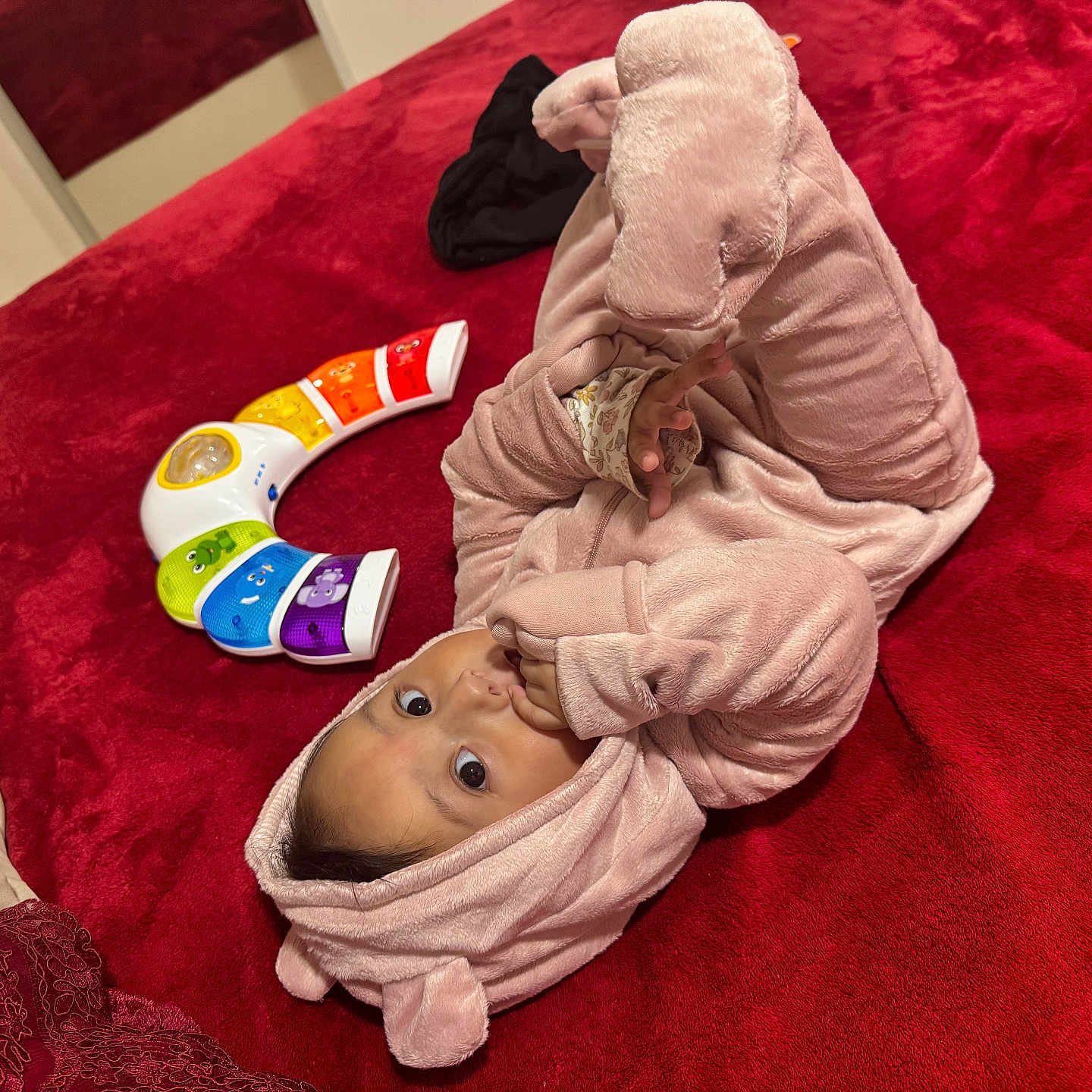 Maelys is registered to the contest to win money with this photo: baby, child, colorful_toy, cozy, curious, cute, face, hand, hood, indoor, infant, lying_down, person, pink_onesie, playful, plush_clothing, red_blanket, soft_texture, sucking_fingers, toy_piano