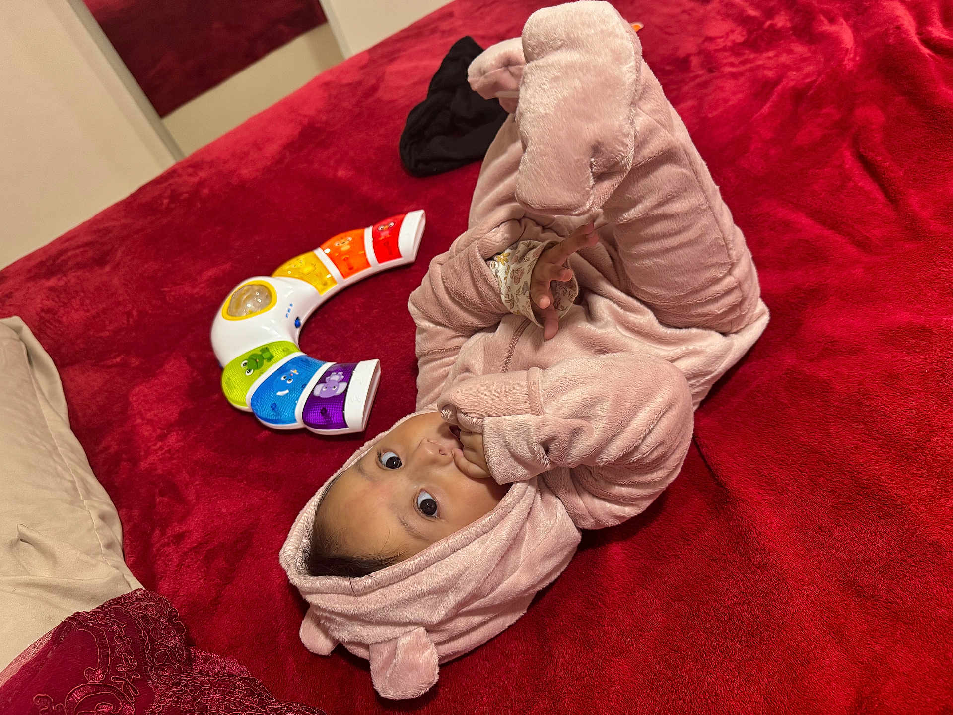 Maelys is registered to the contest to win money with this photo: baby, child, pink_onesie, hood, red_blanket, toy_piano, colorful_toy, lying_down, indoor, curious, sucking_fingers, soft_texture, plush_clothing, infant, cute, playful, person, face, hand, cozy