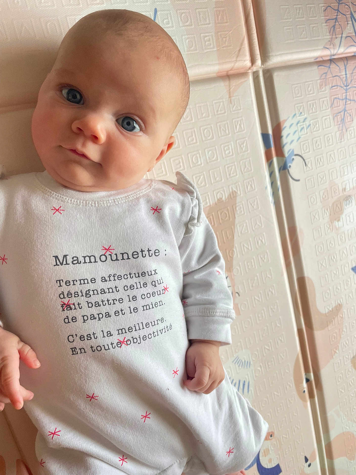 Juliette participe au concours pour gagner de l'argent avec cette photo : baby, infant, face, blue_eyes, white_clothing, text_on_clothing, hand, patterned_background, pastel_colors, cute, portrait, child, soft_lighting, indoor, playful, closeup, young, expression, person, skin