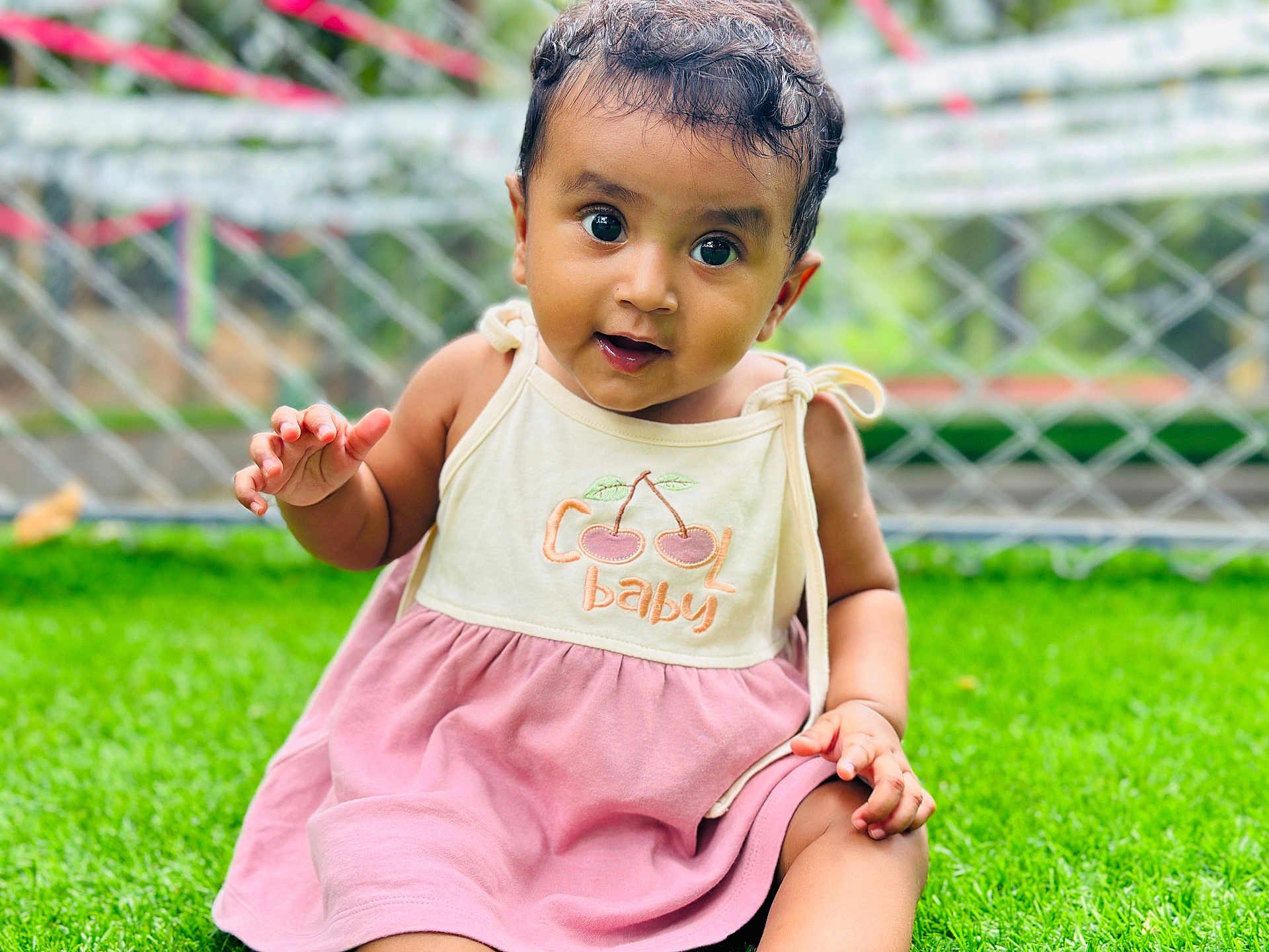 Ashlyn joined the competition — help win amazing prizes! baby, child, grass, outdoor, dress, cute, smiling, face, portrait, sitting, green, fence, person, happy, young_child, curly_hair, nature, summer, playful, eyes