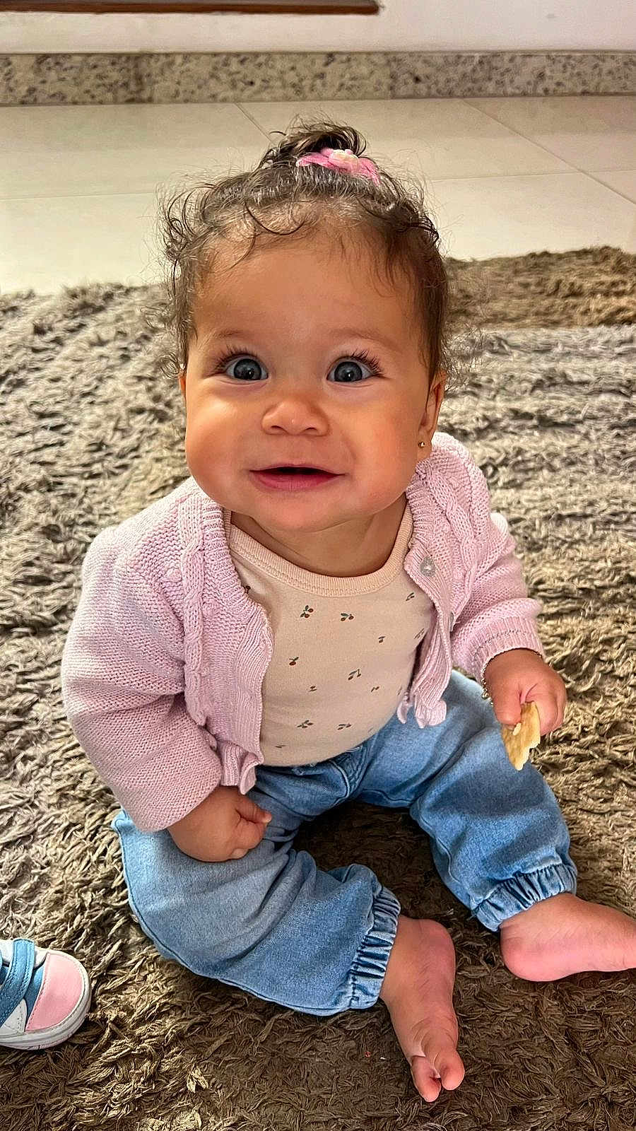 Ayla participe au concours pour gagner de l'argent avec cette photo : baby, child, smiling, sitting, indoor, carpet, pink_sweater, jeans, barefoot, snack, curly_hair, cute, happy, portrait, toddler, playful, small_hair_tie, floor, casual_clothing, bright_eyes