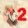 baby, sleeping, flower_headband, white_blanket, pink_blanket, red_roses, number_two, cozy_clothing, infant, cute, portrait, soft_texture, resting, newborn, hands, feet, buttoned_cardigan, patterned_pants, celebration, milestone