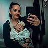 woman, baby, mirror_selfie, smiling, indoor, headband, white_cardigan, floral_pants, holding, portrait, casual_clothing, happy, person, mobile_phone, reflection, cozy, home, cute, mother, child