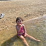child, water, swimsuit, beach, rocks, sunlight, smile, sitting, outdoor, nature, summer, playful, small, person, shallow_water, barefoot, legs, happy, daytime, recreation