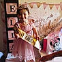 child, birthday, party, dress, tiara, sash, table, balloons, banner, gift_bag, minnie_mouse, pink, smile, celebration, indoor, person, happy, shoes, hand, decorations