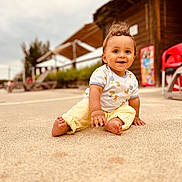 Livio a rejoint le concours — aidez-le/la à gagner de superbes lots ! toddler, child, smiling, outdoor, sand, playful, happy, casual_clothing, yellow_pants, white_shirt, curly_hair, barefoot, wooden_building, red_chair, daylight, nature, portrait, sitting, cute, young_child