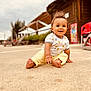 toddler, child, smiling, outdoor, sand, playful, happy, casual_clothing, yellow_pants, white_shirt, curly_hair, barefoot, wooden_building, red_chair, daylight, nature, portrait, sitting, cute, young_child