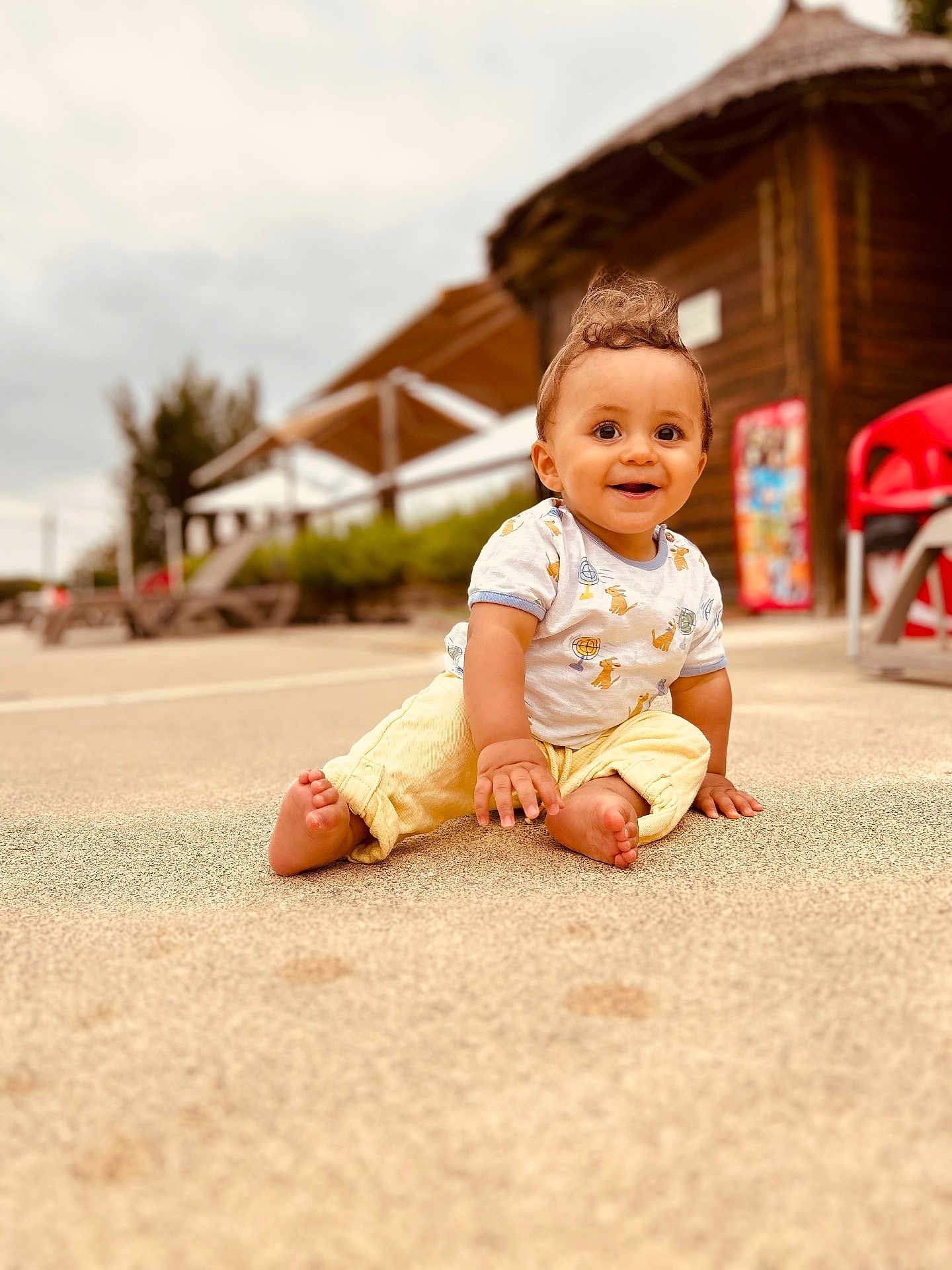 Livio a rejoint le concours — aidez-le/la à gagner de superbes lots ! toddler, child, smiling, outdoor, sand, playful, happy, casual_clothing, yellow_pants, white_shirt, curly_hair, barefoot, wooden_building, red_chair, daylight, nature, portrait, sitting, cute, young_child