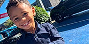 Aron participe au concours pour gagner de l'argent avec cette photo : child, boy, smiling, curly_hair, blue_shirt, outdoor, parking_lot, car, sunny, daylight, happy, person, urban, pavement, casual_clothing, portrait, standing, greenery, metal_structure, blue_sky