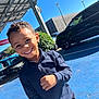 child, boy, smiling, curly_hair, blue_shirt, outdoor, parking_lot, car, sunny, daylight, happy, person, urban, pavement, casual_clothing, portrait, standing, greenery, metal_structure, blue_sky
