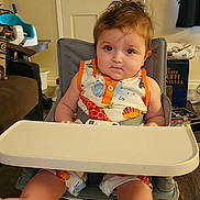 Rafael is registered to the contest to win money with this photo: baby, high_chair, child, indoor, furniture, table, clothing, person, face, foot, hair, expression, living_room, door, curtain, book, paper, toy, floor, seat
