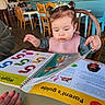 toddler, child, book, reading, parent's_guide, high_chair, hair_buns, pink_bib, table, hand, indoor, wooden_chairs, cafe, curious, learning, colorful, pages, furniture, person, young_child
