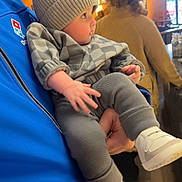 Ace is registered to the contest to win money with this photo: baby, child, person, indoor, hat, clothing, drool, blue_jacket, holding, curious, blurred_background, warm_lighting, flooring, shoes, knit_hat, checkered_outfit, hand, portrait, casual, people