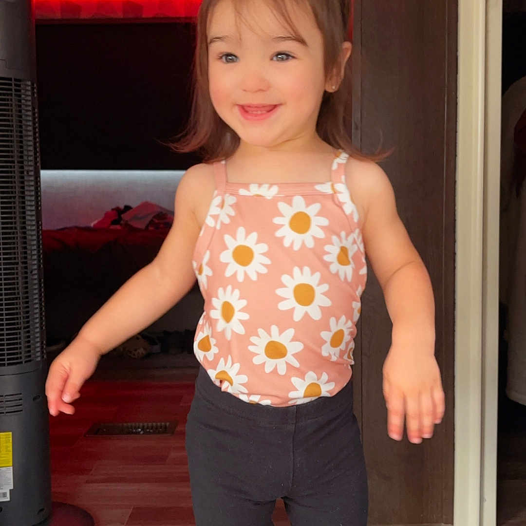 Byntlee is registered to the contest to win money with this photo: black_pants, cabinet, casual_clothing, child, cute, daisy_pattern, doorway, fan, flooring, hair_tied, happy, indoor, person, pink_top, red_led_lights, shoes, smiling, standing, toddler, young_child