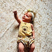 Lainey is registered to the contest to win money with this photo: baby, infant, child, yellow_outfit, headband, sunflower_print, raising_arm, lying_down, patterned_rug, cute, adorable, soft_lighting, indoors, barefoot, smiling, young_child, one_person, portrait, casual_clothing, relaxed