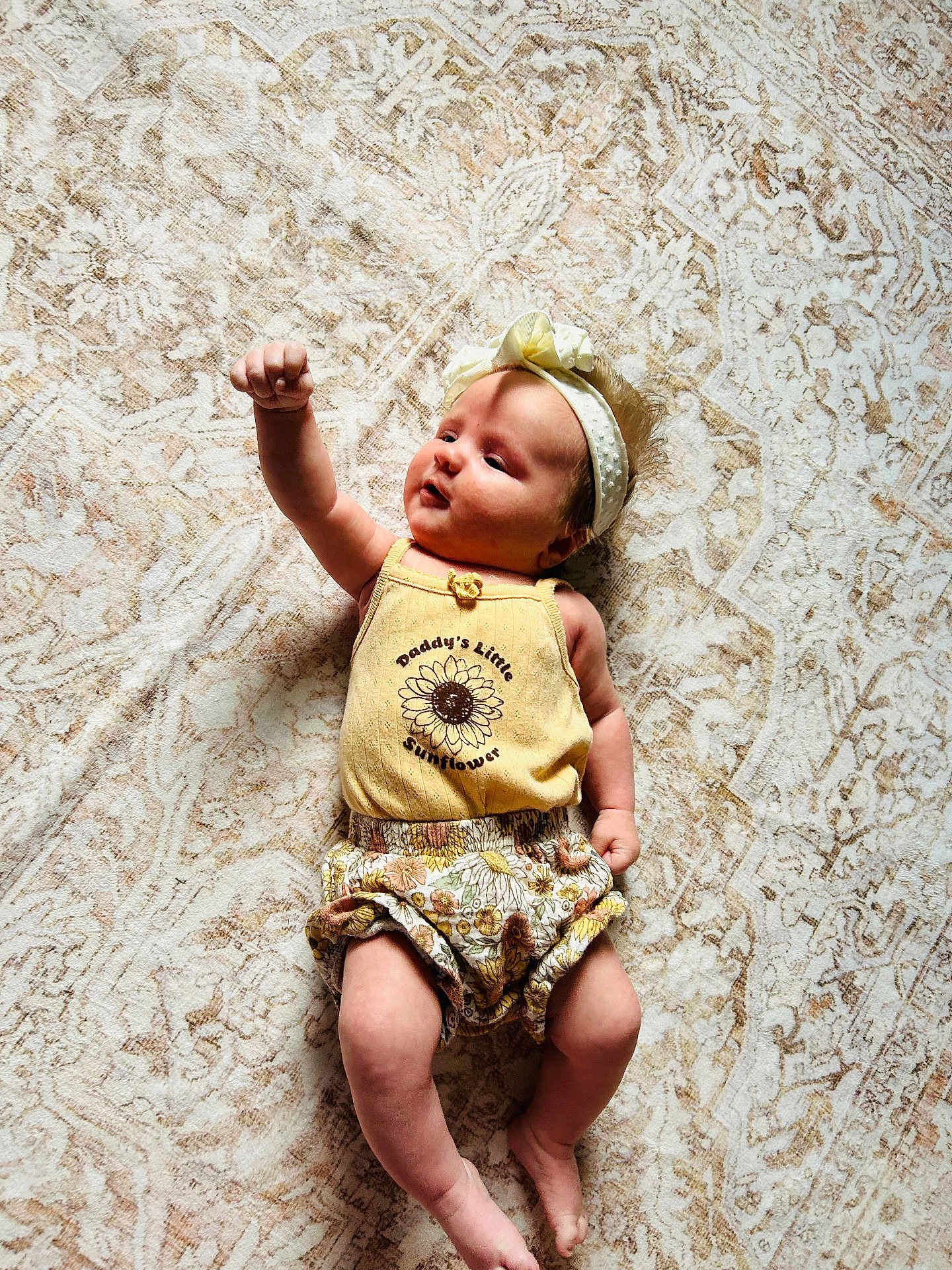 Lainey is registered to the contest to win money with this photo: baby, infant, child, yellow_outfit, headband, sunflower_print, raising_arm, lying_down, patterned_rug, cute, adorable, soft_lighting, indoors, barefoot, smiling, young_child, one_person, portrait, casual_clothing, relaxed