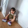 toddler, child, curly_hair, green_dress, sneakers, wooden_floor, white_wall, indoor, sitting, floor, shoes, baby, cute, portrait, casual_clothing, curious, young_child, person, home, expression