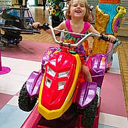 Erin a rejoint le concours — aidez-le/la à gagner de superbes lots ! child, girl, colorful_dress, kiddie_ride, atv_ride, indoor, shopping_mall, arcade_machine, statue, playful, joyful, smiling, toy, floor_tiles, handlebars, people_in_background, plant, sign, casual_clothing, fun