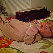 Oakley is registered to the contest to win money with this photo: baby, infant, pacifier, onesie, bed, blanket, quilt, floral_pattern, sleepwear, lying_down, indoor, child, person, bedroom, soft_lighting, curious_expression, home, cuddly, resting, cozy