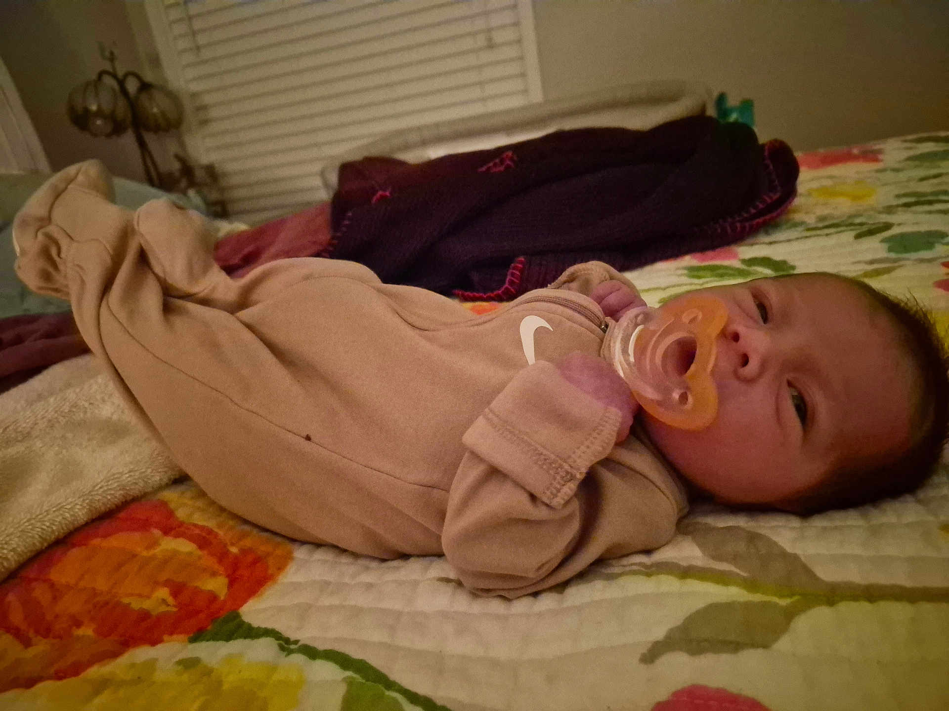 Oakley is registered to the contest to win money with this photo: baby, infant, pacifier, onesie, bed, blanket, quilt, floral_pattern, sleepwear, lying_down, indoor, child, person, bedroom, soft_lighting, curious_expression, home, cuddly, resting, cozy