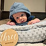 Lincoln is registered to the contest to win money with this photo: baby, infant, hood, blue_clothing, blanket, cushion, wooden_sign, milestone, three_months, bed, cute, child, portrait, wide_eyes, soft_texture, indoor, resting, face, hand, young_baby