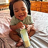 baby, bottle, milk, bed, smiling, happy, indoor, wooden_furniture, window, air_conditioner, clothing, head_covering, person, infant, human, furniture, room, dressing_table, curtain, skin