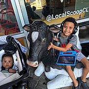 Seth is registered to the contest to win money with this photo: child, baby, stroller, headphones, tablet, cow_statue, bench, smile, outdoor, window, reflection, seat, boy, toy, casual_clothing, happy, urban, sidewalk, playful, technology