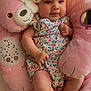 baby, baby_outfit, cherry_pattern, child, colorful_clothing, cozy, cute, flower, headband, indoors, infant, pink_cushion, plush_toy, portrait, relaxed, smiling, soft_toys, star_pattern, teddy_bear, yellow_bow