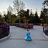 child, girl, princess_dress, toy, stuffed_animal, park, trees, lights, pathway, purple_railings, evening, outdoor, nature, pavement, lanterns, costume, young_child, playful, twilight, scenic