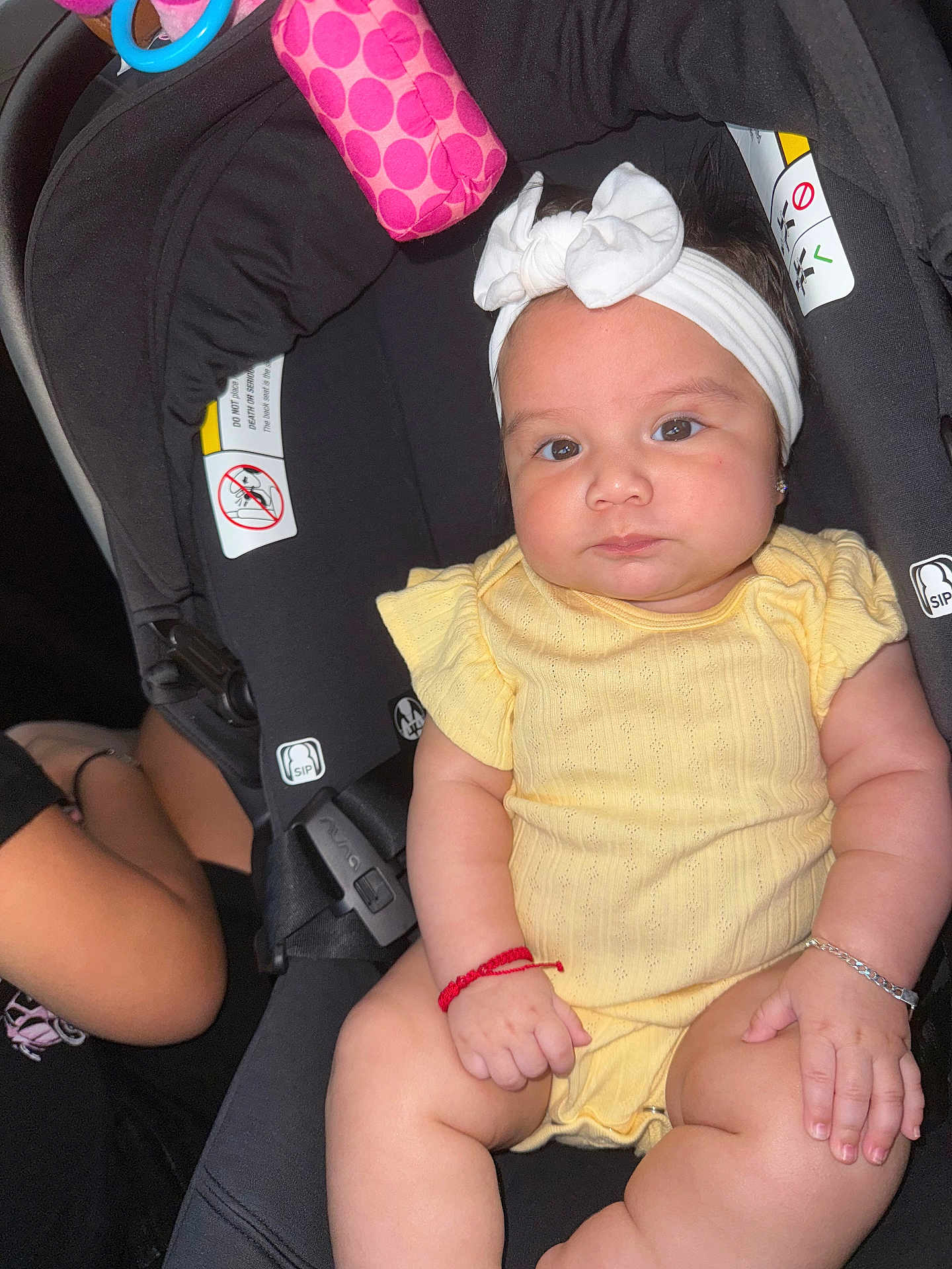 Brianna is registered to the contest to win money with this photo: baby, child, car_seat, yellow_clothing, headband, bracelet, infant, chubby, cute, indoor, person, seatbelt, black_seat, pink_toy, expressionless, wrists, legs, skin, closeup, portrait