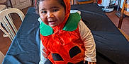 Enëa a rejoint le concours — aidez-le/la à gagner de superbes lots ! black_tablecloth, chair, child, costume, cozy, curly_hair, curtains, furniture, happy, home, indoor, orange, person, playful, pumpkin_costume, smiling, socks, table, toddler, window