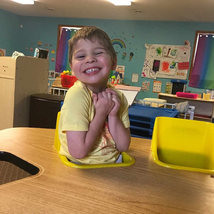 Armani joined the competition — help win amazing prizes! artwork, child, classroom, colorful, education, furniture, hands_clasped, happy, indoor, joyful, kid, person, playroom, seat, smiling, table, wall_decor, yellow_chair, yellow_shirt, young_child