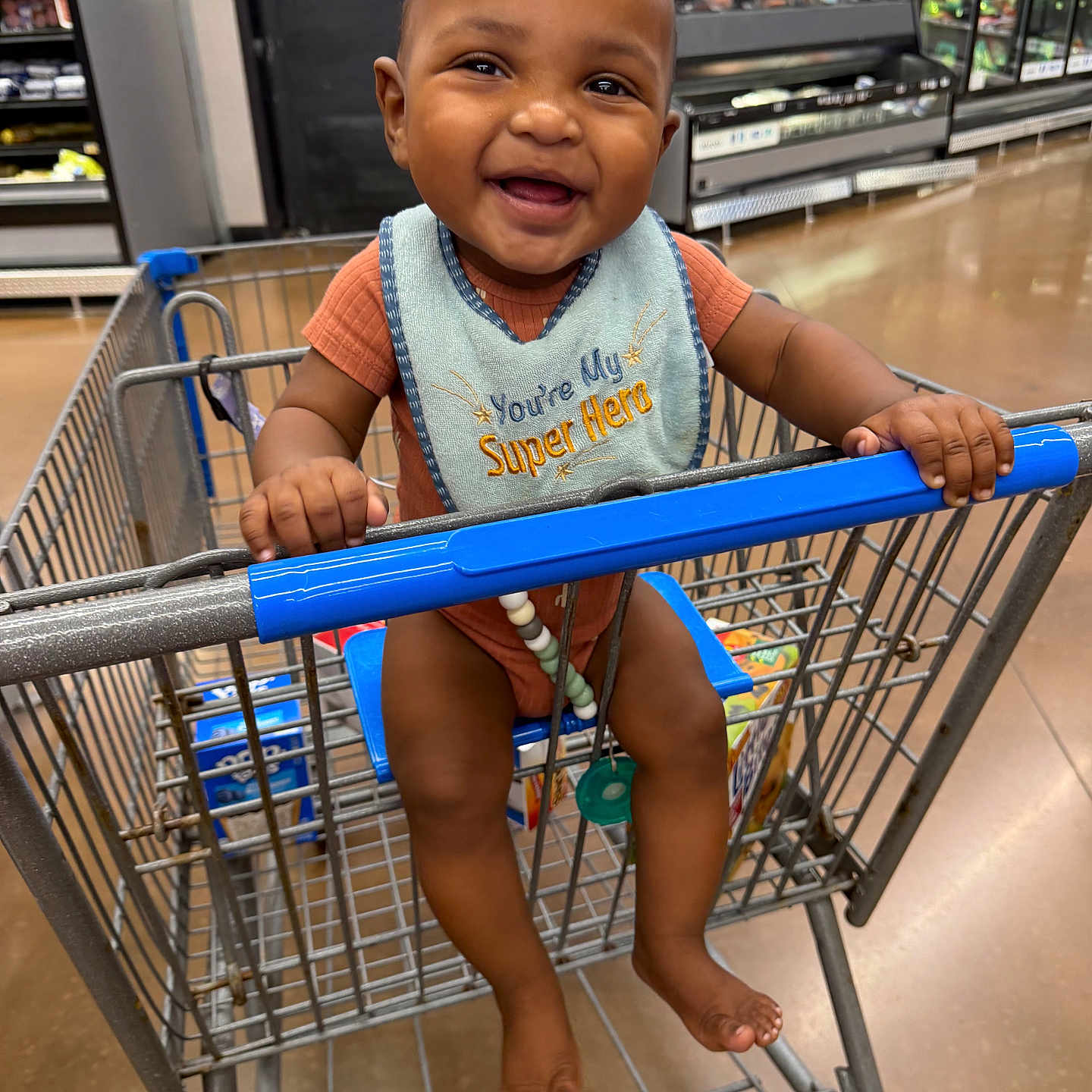 Zymir is registered to the contest to win money with this photo: baby, shopping_cart, grocery_store, smiling, child, bib, happy, indoor, person, holding, clothing, seat, footwear_free, retail, shopping, super_hero, infant, store_shelves, floor, metal