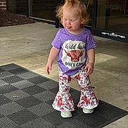 Luna is registered to the contest to win money with this photo: toddler, child, purple_shirt, graphic_tshirt, floral_pants, bell_bottoms, topknot_hairstyle, tongue_out, black_mat, concrete_floor, entrance, glass_door, brick_wall, outdoor, playful_expression, casual_clothing, daylight, person, standing, cute