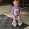 toddler, child, purple_shirt, graphic_tshirt, floral_pants, bell_bottoms, topknot_hairstyle, tongue_out, black_mat, concrete_floor, entrance, glass_door, brick_wall, outdoor, playful_expression, casual_clothing, daylight, person, standing, cute