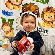 Mylan a rejoint le concours — aidez-le/la à gagner de superbes lots ! baby, child, toy_truck, panda_onesie, blanket, lion_illustration, stuffed_toy, cushion, smile, indoor, cute, happy, playful, clothing, person, portrait, infant, soft_toy, colorful, seated